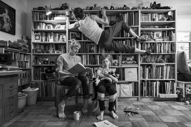 A black and white photo shows a man climbing a bookcase, a woman reading and a child holding a cat.